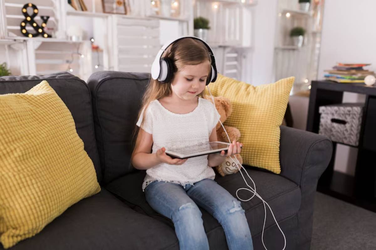 A child with long hair sits on a gray couch, wearing headphones and holding a tablet, surrounded by yellow pillows and a teddy bear.