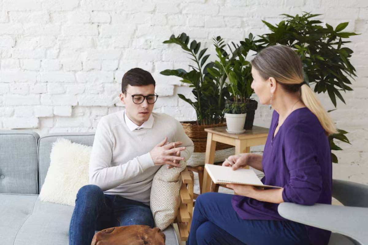 A man sits on a couch, engaged in conversation with a woman holding a notebook, surrounded by indoor plants and a cozy setting.