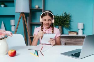 A young girl wearing headphones sits at a desk with a laptop, colored pencils, and a notepad, smiling as she writes and follows along in a virtual homeschool session, set in a brightly lit, organized study space.