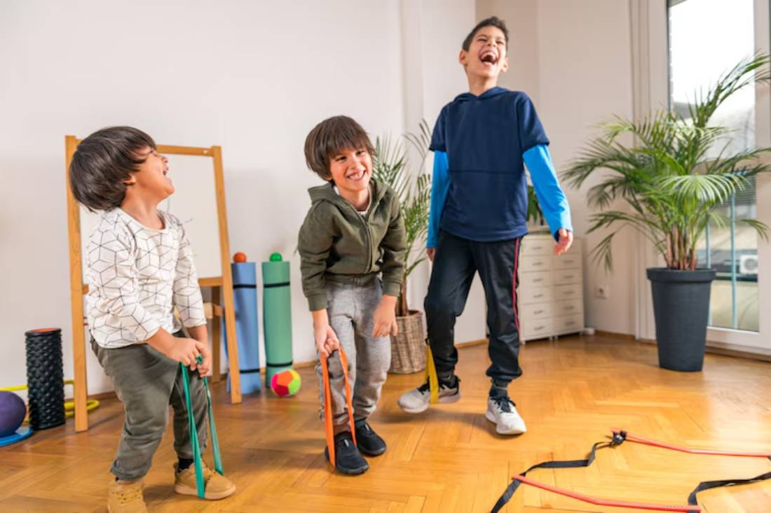 Three children engage in playful activity with rubber bands in a bright, airy room filled with colorful toys and plants.