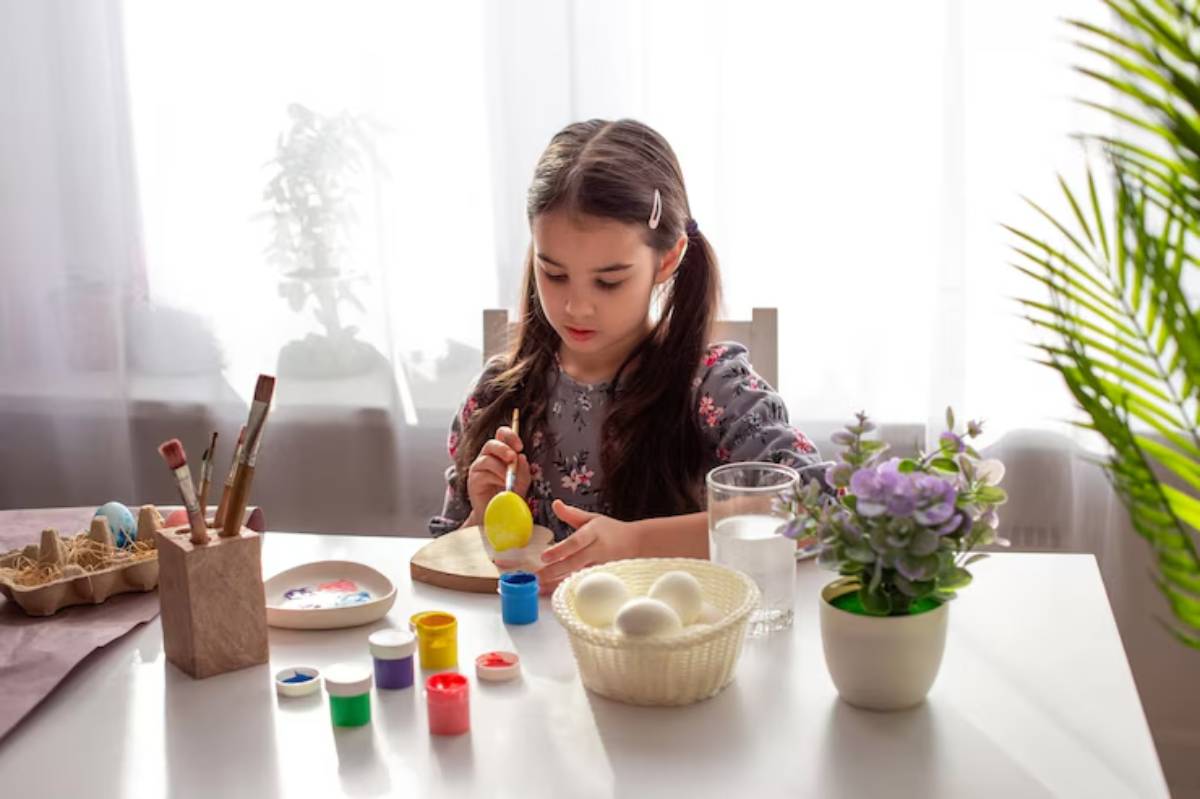 A child paints an egg at a table, surrounded by colorful paints, a glass of water, and a vase of flowers in a bright room.