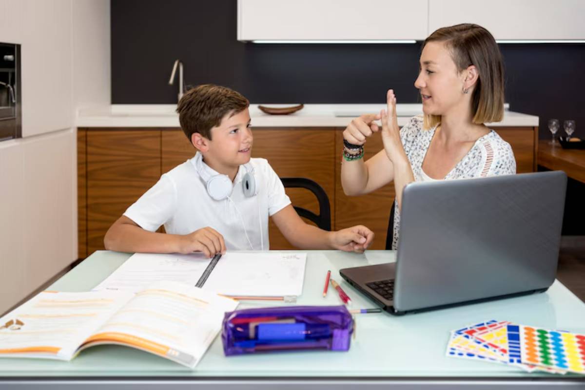 A woman teaches a boy at a modern kitchen table, surrounded by school supplies and a laptop, fostering a learning environment.