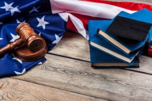 A wooden gavel rests on an American flag, next to stacked blue books and a graduation cap, symbolizing law and education.