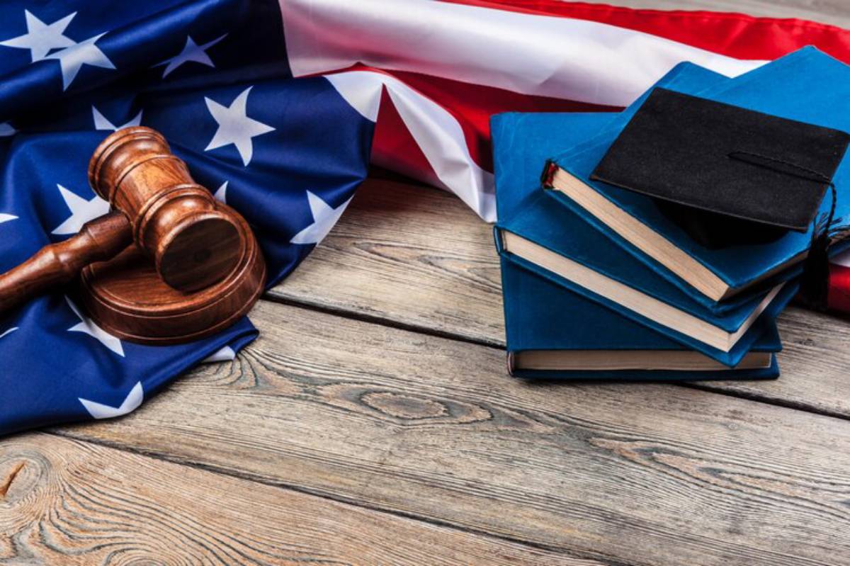 A wooden gavel rests on an American flag, next to stacked blue books and a graduation cap, symbolizing law and education.