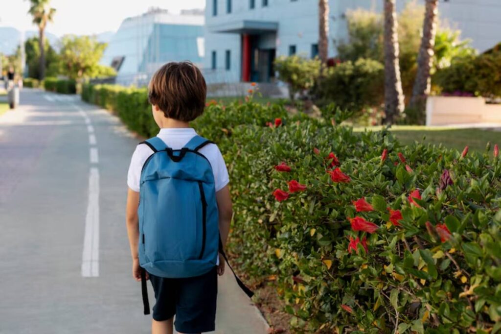 A young boy in a blue backpack walks along a sunny path beside flowering bushes, heading towards a modern building.