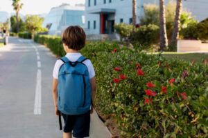 A young boy in a blue backpack walks along a sunny path beside flowering bushes, heading towards a modern building.