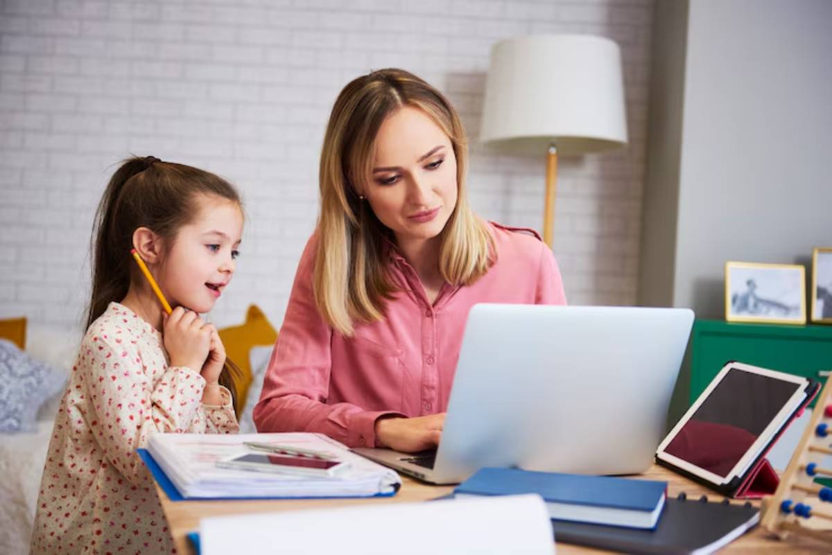 A woman and a child engage in remote learning at a table with a laptop, books, and stationery in a cozy, brightly lit room.