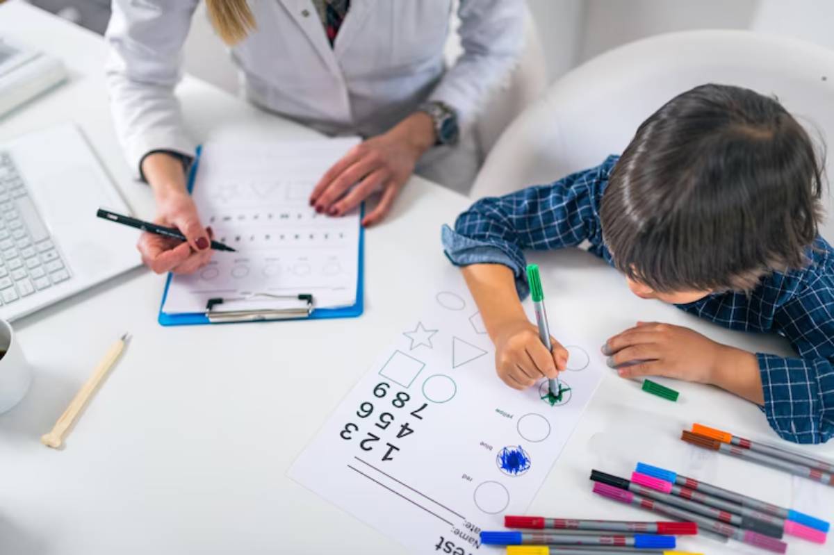 A child in a checkered shirt colors a worksheet while a woman notes on a clipboard, surrounded by markers and a laptop.