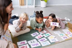 A group of young children and a teacher engaging in a playful learning activity on the floor, using colorful alphabet flashcards to practice letter recognition in a bright and friendly classroom setting.