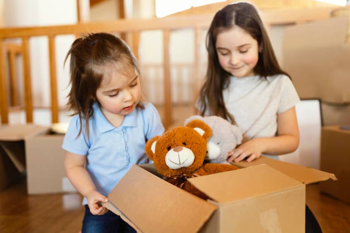 Two children unbox stuffed animals, including a brown teddy bear and a gray plush, amidst moving boxes in a cozy room.