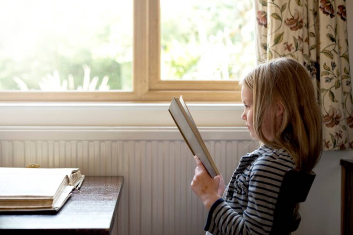 A child sitting by a sunlit window, engrossed in reading a book, surrounded by a warm, inviting room with floral curtains.