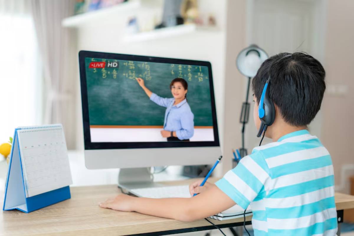 A child in a striped shirt listens to a live online lesson, taking notes while seated at a desk with a calendar and stationery.