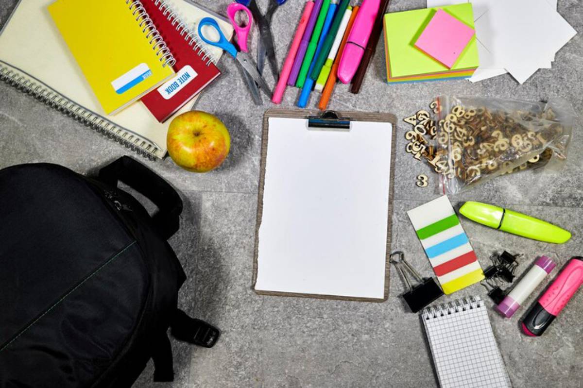 Flat lay of school supplies, including notebooks, pens, sticky notes, an apple, and a backpack, arranged on a textured surface.