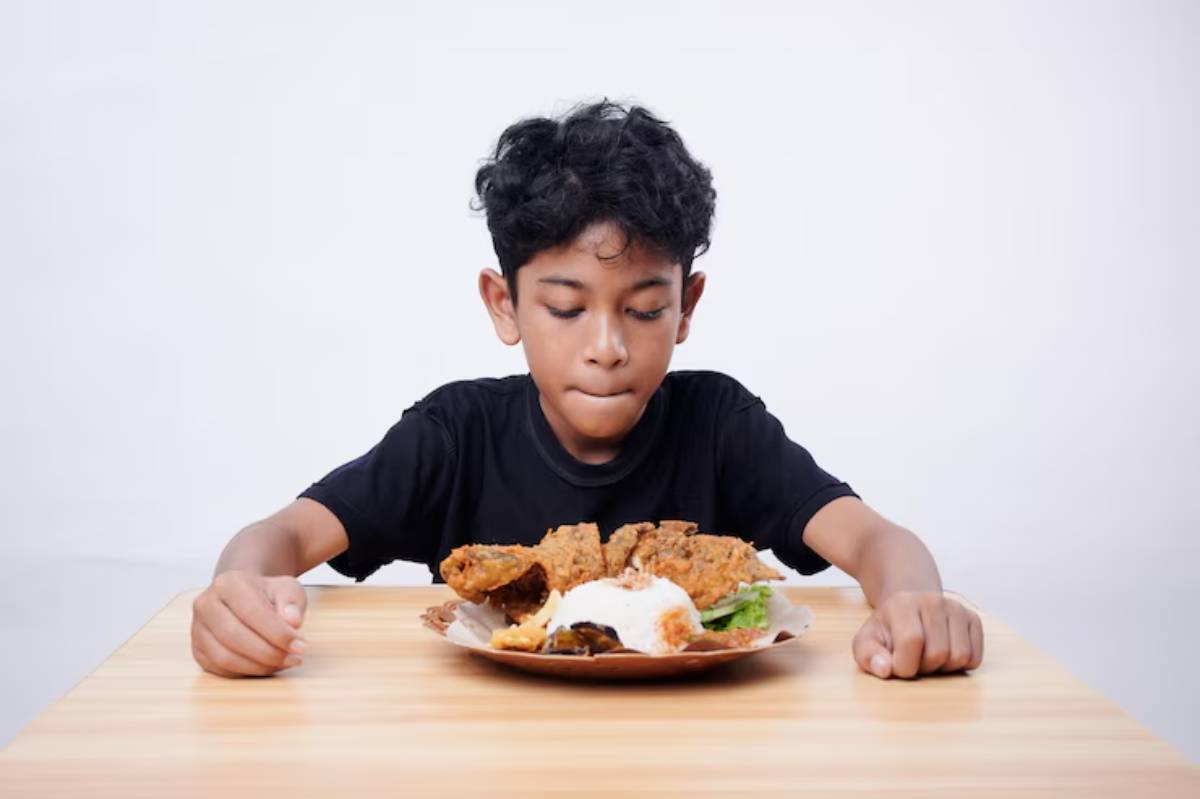 A child sits at a table with a plate of fried chicken, rice, and vegetables, eagerly waiting to eat.