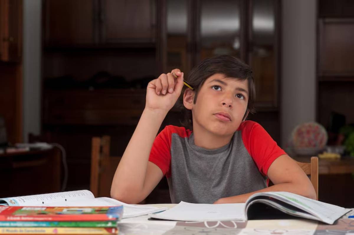 A child sits at a table covered with books, holding a pencil thoughtfully, surrounded by a cozy, indoor setting.