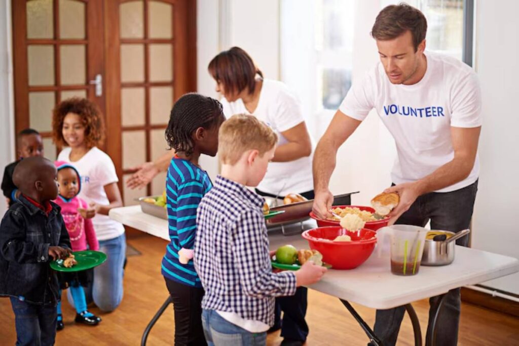 Volunteers serve food to children at a table, showcasing a vibrant community meal setting with plates of food and enthusiastic faces.