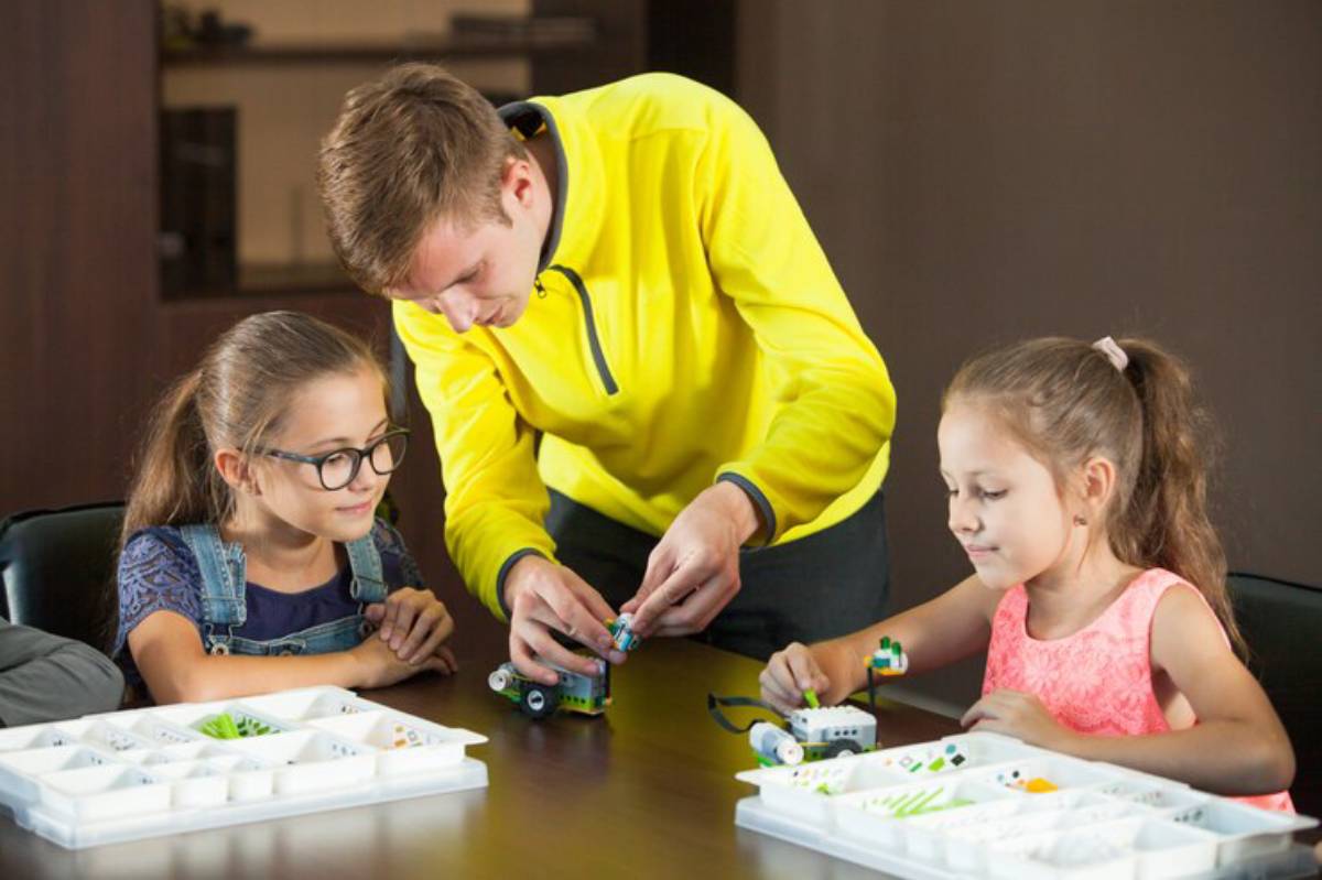A person helps two children build small robotic models using colorful pieces on a table filled with various components.