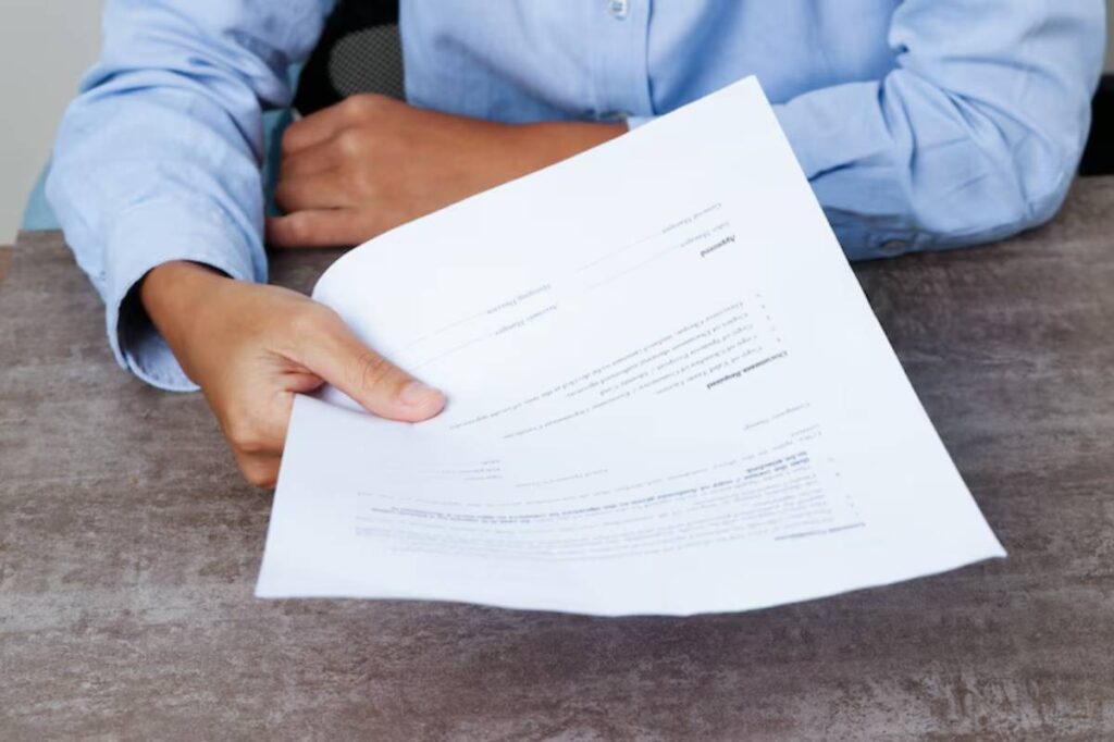 A person in a light blue shirt is handing a document across a wooden table, showcasing a partially visible printed page.