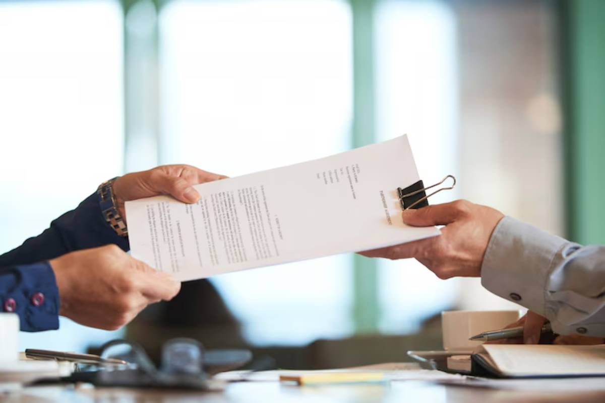 Two hands exchanging a document with a binder clip, set on a table with office supplies and a cup in the background.