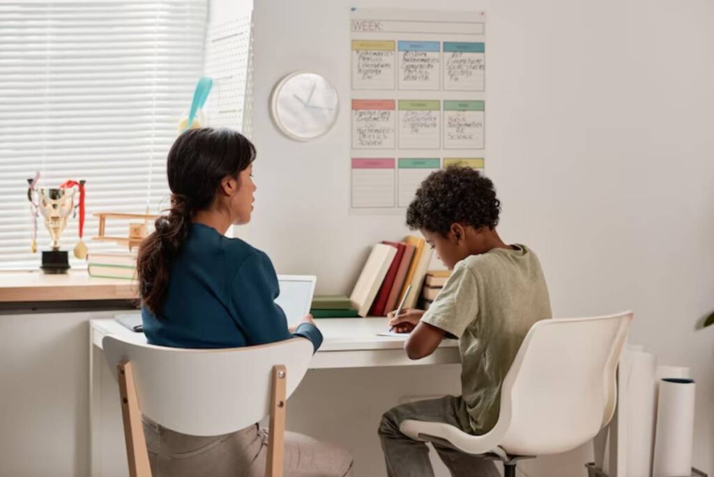 A student sits at a desk writing while a tutor assists from the opposite side, with a calendar and books visible in the background.