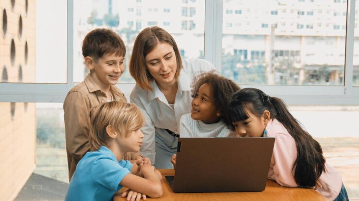 A group of children gathered around a laptop, engaged in learning, with a bright window view behind them.