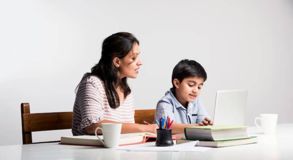A woman and a child sit at a table with books, stationery, and a laptop, engaged in study or learning together.