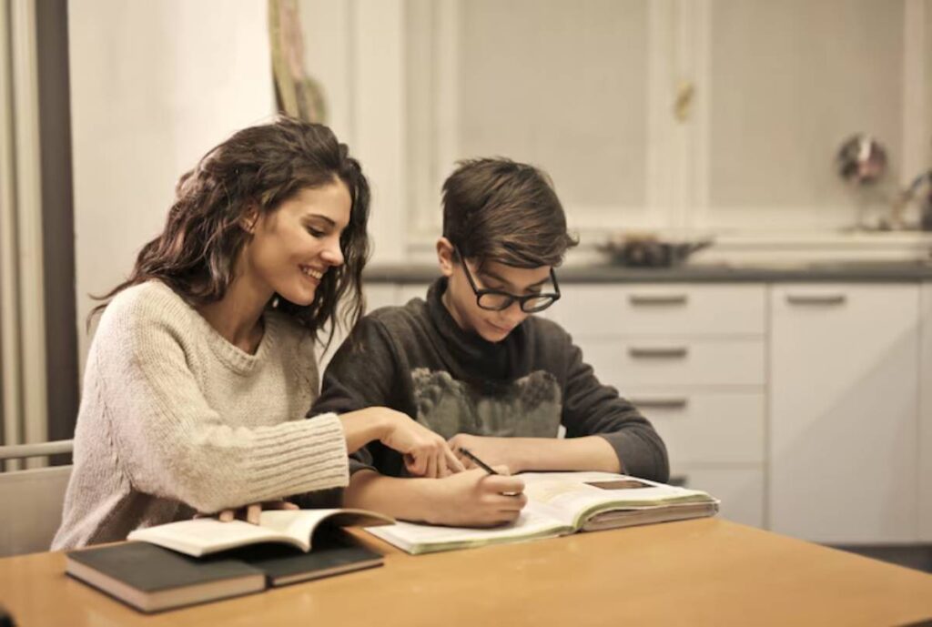 A person in a sweater points at a book, assisting another individual with studying at a table filled with open textbooks.