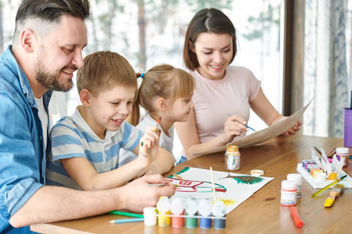 A family enjoys a creative art session at a table, surrounded by paints, brushes, and colorful drawings in a bright, airy room.