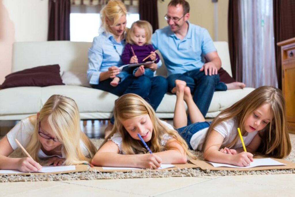 Three young girls lie on the floor, drawing on paper, while a family sits on a couch in the background, enjoying time together.