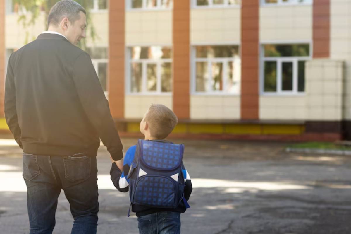 A parent and child walk hand in hand towards a school building, with the child carrying a blue backpack.