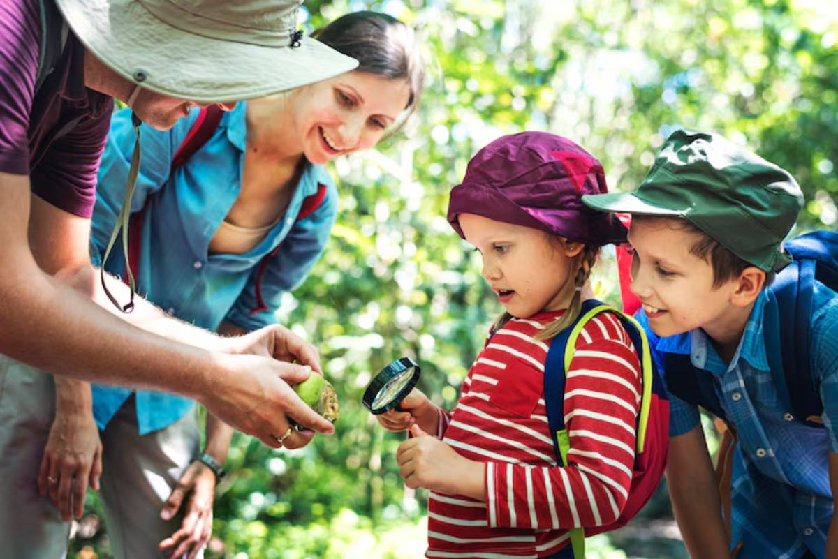 A group of children and adults explore nature, closely observing a green object held by an adult while one uses a magnifying glass.