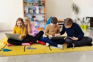 A group of children engaged in a digital learning session, sitting on a colorful rug with laptops and a tablet in a bright room.