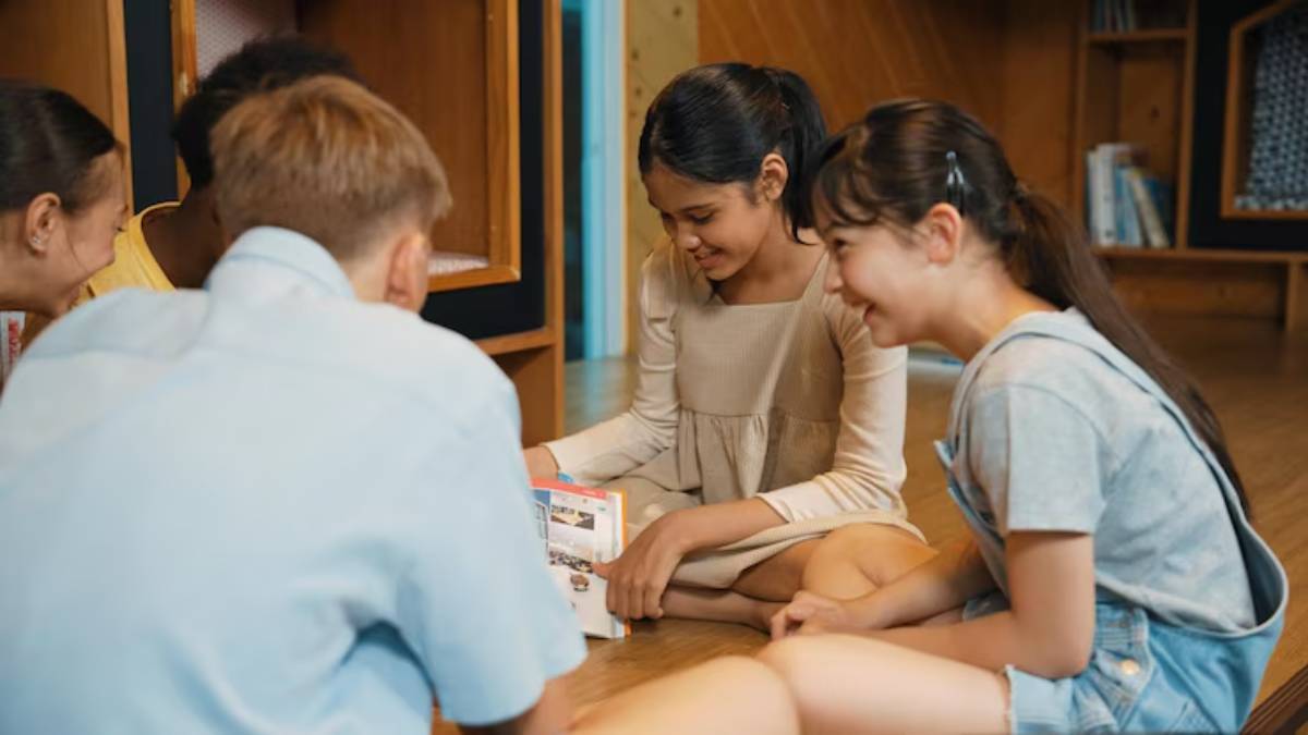 A group of young people sitting on the floor, intently reading a book together in a cozy indoor space with wooden shelves.