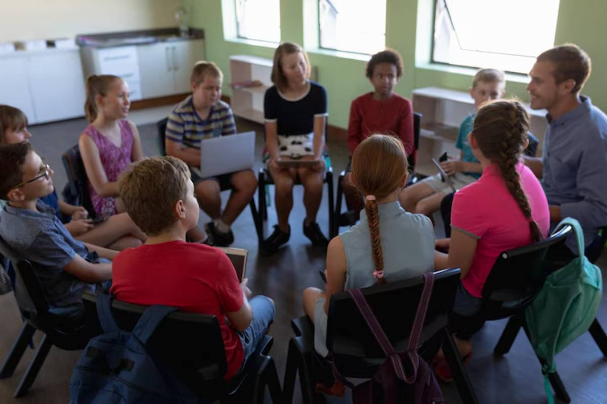 A diverse group of children sits in a circle on chairs, engaged in discussion, in a bright, modern classroom setting.