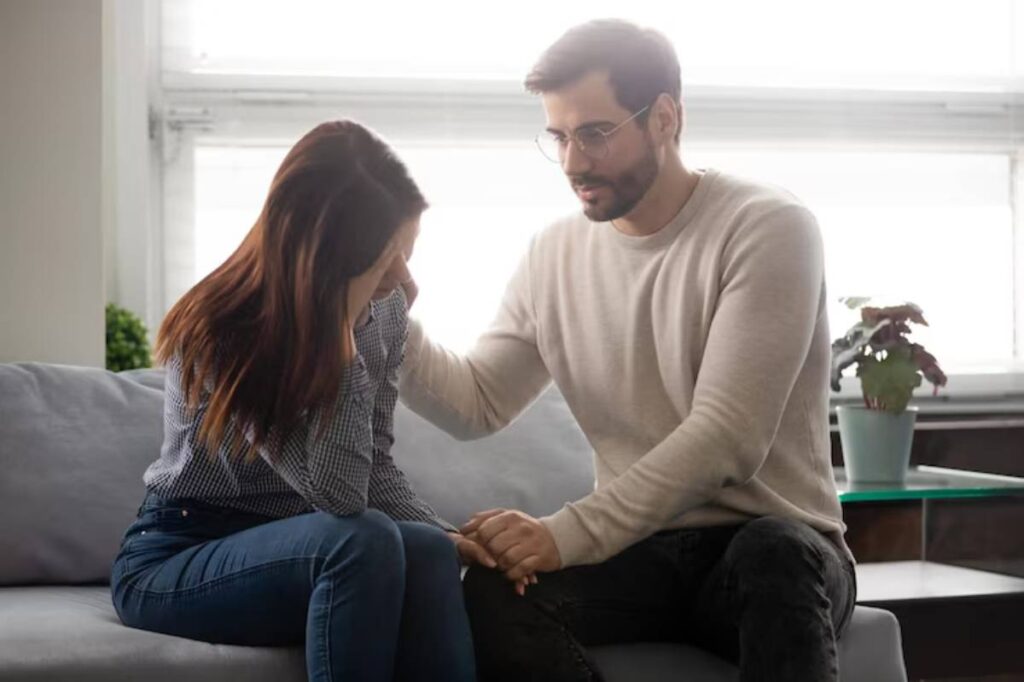 A man comforts a woman sitting on a couch, displaying a supportive and caring interaction in a cozy room.