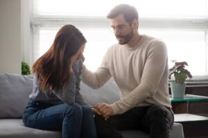 A man comforts a woman sitting on a couch, displaying a supportive and caring interaction in a cozy room.