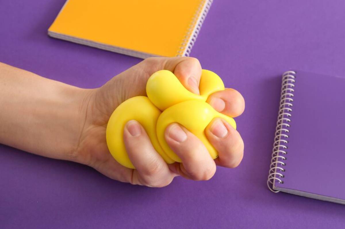 A hand squeezes a cluster of yellow stress balls against a purple background, with orange and purple notebooks nearby.