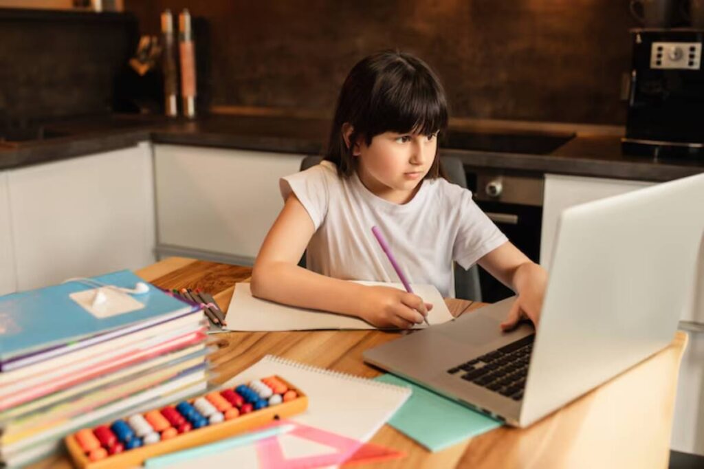 A child in a white t-shirt sits at a wooden table, writing on paper while working on a laptop surrounded by school supplies.