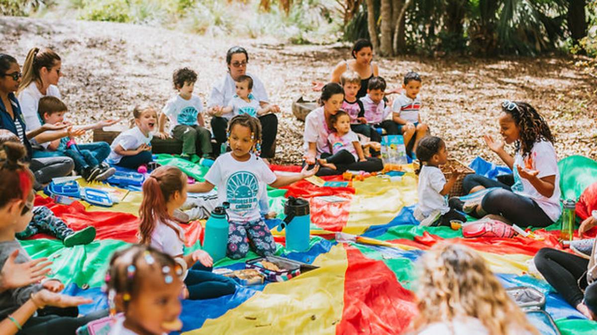 A diverse group of children and adults engage in activities on a colorful blanket in a vibrant outdoor setting.