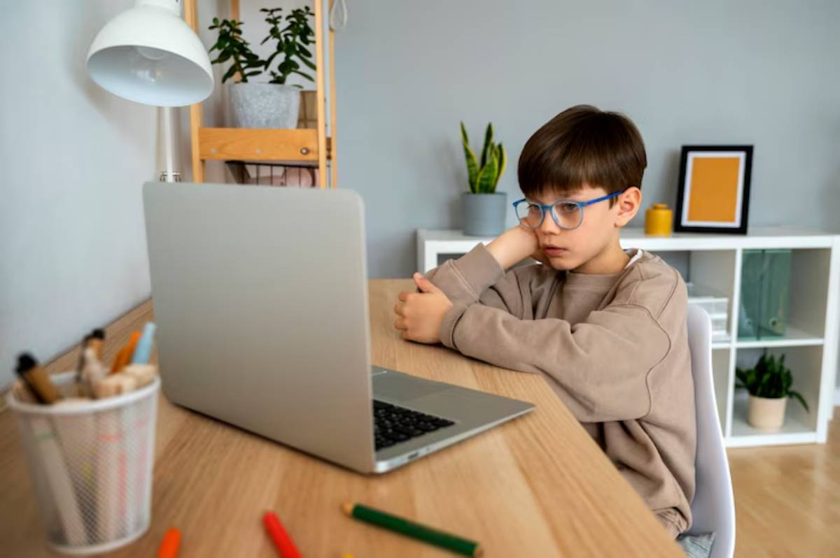 A child sits at a wooden desk, gazing at a laptop, with plants and stationery nearby, in a cozy, modern workspace.