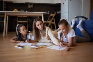 A teacher and two children are lying on the floor, engaged in painting with colorful watercolor palettes in a cozy room.