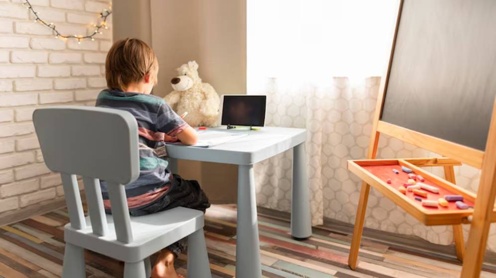 A child sits at a small desk with a tablet and notebook, facing a blackboard and a teddy bear in a cozy, well-lit room.