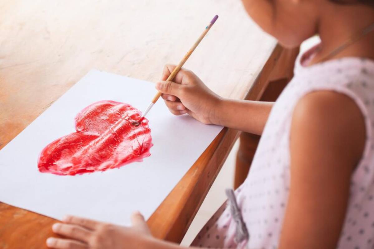 A child paints a bright red heart on white paper, holding a brush, focused on their artwork, with a wooden table in the background.