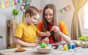 A child in a yellow shirt and an adult in an orange shirt decorate Easter eggs with paint at a table filled with art supplies.