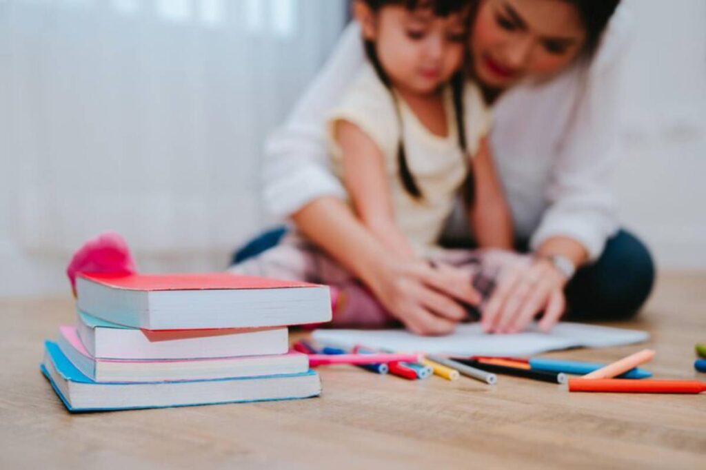 A stack of colorful books and various markers scattered on a wooden floor, with a child working on a drawing alongside an adult.