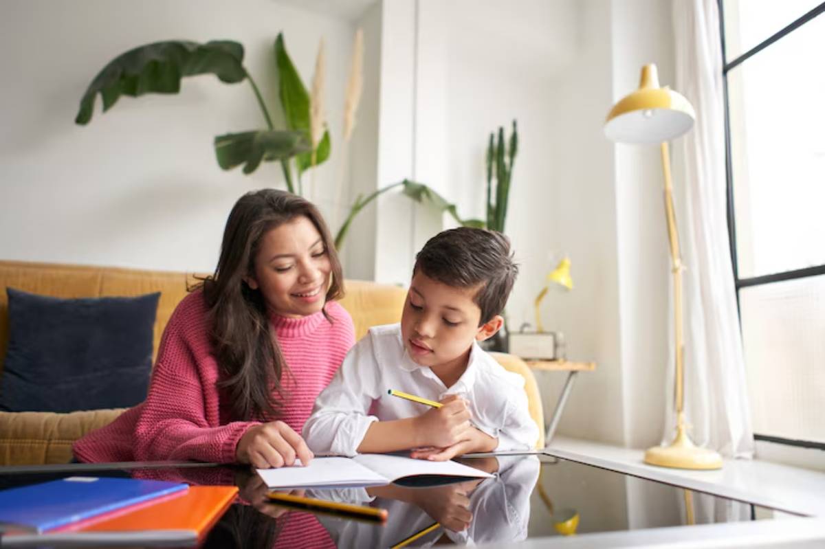 A young girl in a pink sweater and a boy in a white shirt work together at a desk, with vibrant plants and natural light in the background.