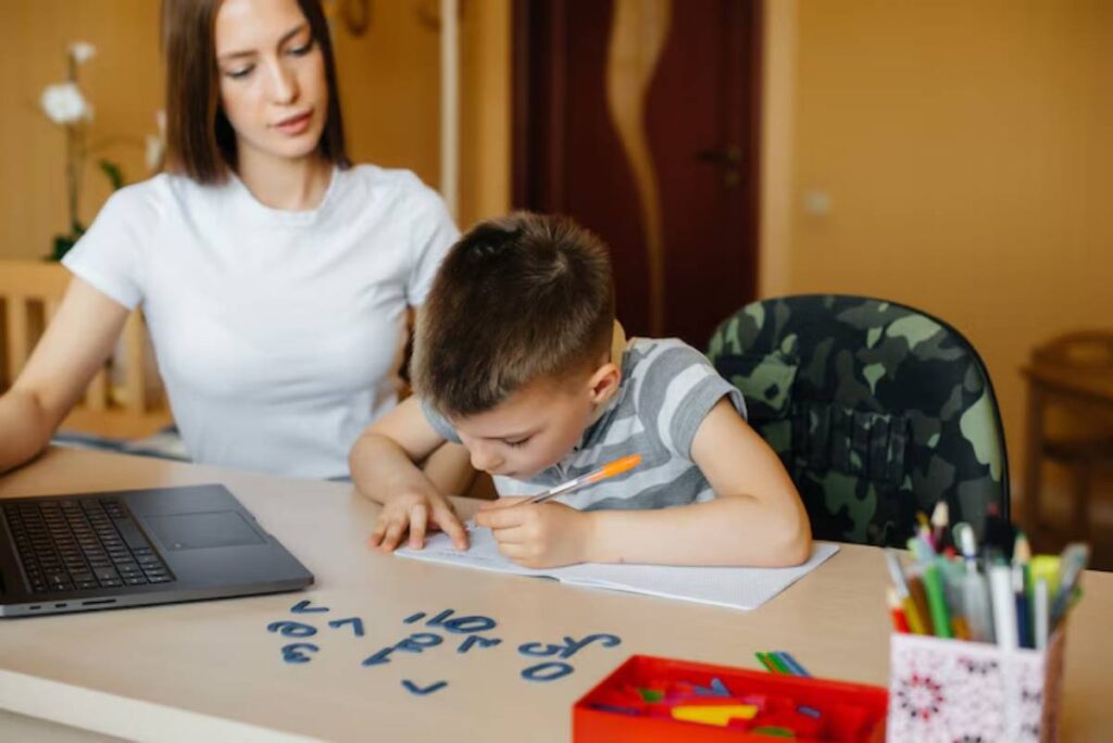 A child diligently writes on paper at a table with a laptop, colorful markers, and geometric shapes scattered around.