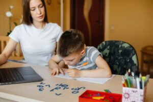 A child diligently writes on paper at a table with a laptop, colorful markers, and geometric shapes scattered around.