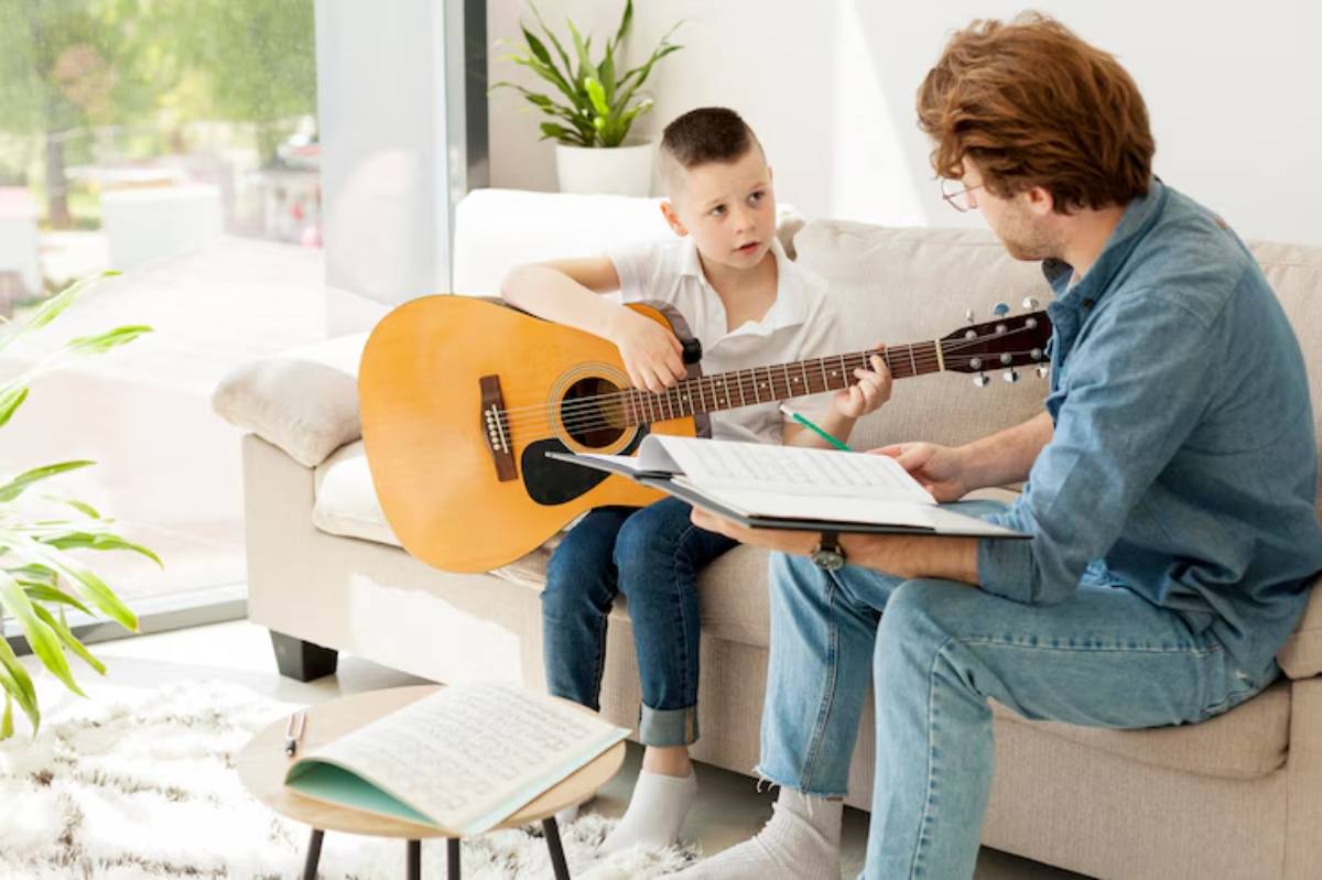 A child plays guitar while sitting on a couch, receiving guidance from an adult with musical notes on a stand nearby.