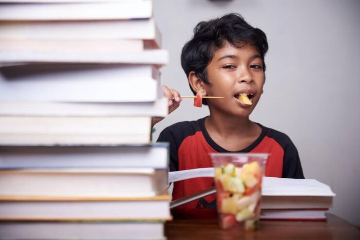 A child in a red t-shirt sits between tall stacks of books, enjoying a colorful cup of fruit.